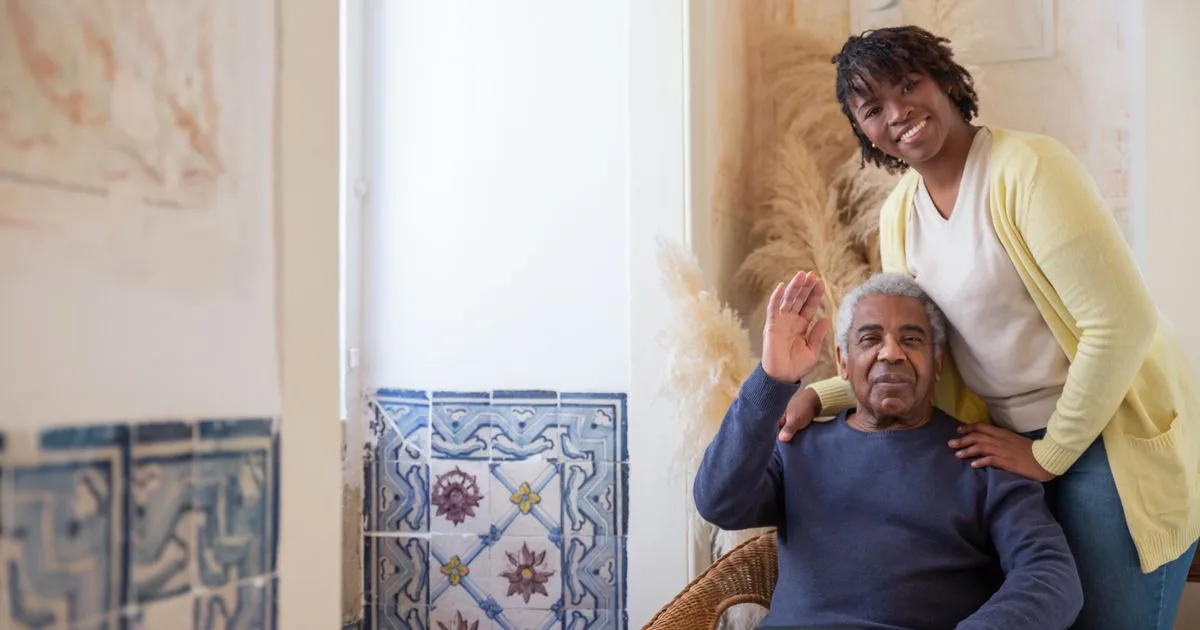 An adult daughter stands with a hand on her seated elderly father's shoulder, both smiling warmly together at home