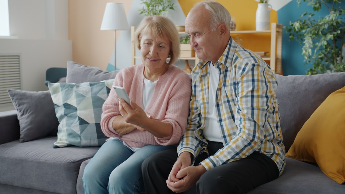 Zwei ältere Menschen entspannen auf einem Sofa und schauen gemeinsam gemütlich auf ein Smartphone